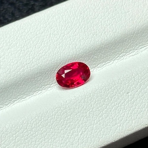 May include: A vibrant, oval-cut ruby gemstone. The gem is a deep, saturated red with facets that catch the light. The ruby is set against a white, textured surface, highlighting its color and brilliance.