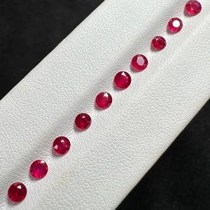 Pode incluir: Uma coleção de rubis redondos, facetados, de cor vermelha profunda, dispostos diagonalmente sobre uma superfície branca. As pedras preciosas são espaçadas uniformemente, mostrando sua lapidação brilhante e cor rica. Os rubis são colocados sobre um fundo preto contrastante.