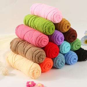 May include: A selection of colourful yarn skeins in shades of pink, green, brown, and blue, arranged in a pyramid. The tightly wound yarn suggests it's suitable for knitting or crochet. The background is a soft, neutral colour, with a few decorative elements.
