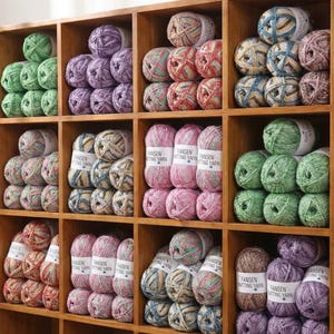 Op de afbeelding: Een houten plank gevuld met diverse kleurrijke bollen wol. De wol is in nette stapels gerangschikt in de vakken. De kleuren van de wol zijn groen, paars, roze en veelkleurige bollen. Elke bol heeft een wit label met "FANSEN KNITTING YARN".