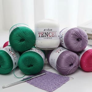 Puede incluir: Una colección de madejas de hilo en varios colores, incluyendo verde, morado y rosa, con el texto "TENCEL YARNS" visible en las etiquetas. También hay un ganchillo y un pequeño cuadrado de lavanda tejido. El hilo está dispuesto sobre una superficie blanca.