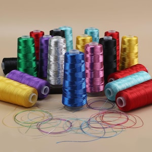 Puede incluir: Una colección de coloridas bobinas de hilo en varios tonos, incluyendo verde, rojo, azul, amarillo, morado y plateado. Los hilos están cuidadosamente enrollados en bobinas cilíndricas, con hebras sueltas en primer plano. Ideal para proyectos de costura y manualidades.