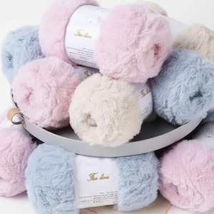 Könnte beinhalten: Eine Ansammlung flauschiger Wollknäuel in Pastellfarben, darunter Rosa, Blau und Creme, angeordnet in einem grauen Tablett. Jedes Knäuel hat ein Etikett mit goldenem Text. Das Garn wirkt weich und strukturiert, geeignet für Strick- oder Häkelprojekte.
