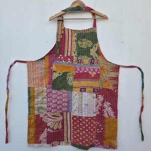 May include: A patchwork apron with a variety of colorful fabric squares in shades of red, green, pink, and yellow. The apron is hanging on a wooden hanger against a white wall. The apron has long ties and a neck strap.