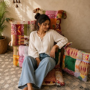 May include: A white long-sleeved blouse with a front tie closure and vertical pleats, paired with light blue denim jeans. The outfit is shown against a backdrop of several square floor cushions featuring a colorful patchwork pattern.