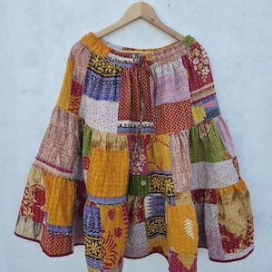 May include: A patchwork skirt with a tiered design, featuring a variety of colorful fabric patterns. The skirt is suspended from a wooden hanger against a plain white background. The skirt's colors include yellow, red, purple, and green.