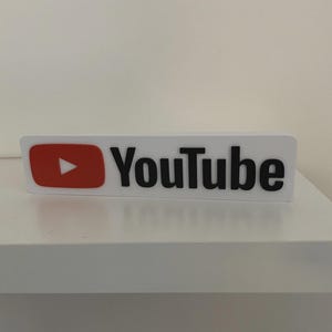 May include: A white rectangular sign displaying the YouTube logo. The logo includes a red square with a white play button and the word "YouTube" in black, sans-serif font. The sign is placed on a white shelf.