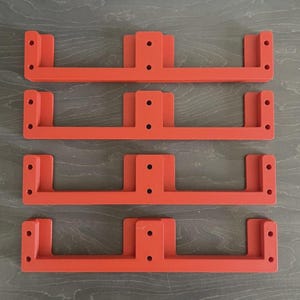 May include: Four bright orange, U-shaped brackets with multiple pre-drilled holes for mounting. The brackets are arranged in a stack, with a simple, functional design. The background is a grey wooden surface.