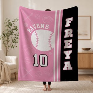 May include: A pink and black fleece blanket featuring a large white baseball graphic, the text RAVENS, the number 10, and the name FREYA in large white block letters. The pink section includes a repeating pattern of baseballs, bats, and gloves.