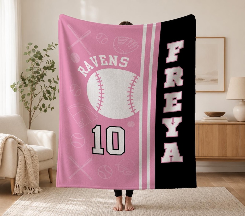 May include: A pink and black fleece blanket featuring a large white baseball graphic, the text RAVENS, the number 10, and the name FREYA in large white block letters. The pink section includes a repeating pattern of baseballs, bats, and gloves.