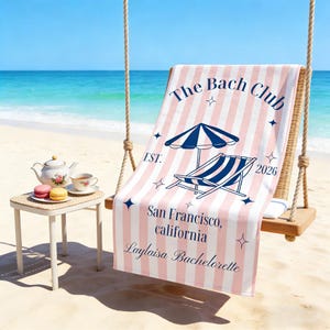 May include: A pink and white striped beach towel with the text "The Bach Club" and "San Francisco, California". The towel features an illustration of a beach umbrella and a lounge chair. A small table with a teapot, teacup, and macarons is in the foreground.