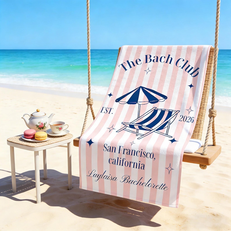 May include: A pink and white striped beach towel with the text "The Bach Club" and "San Francisco, California". The towel features an illustration of a beach umbrella and a lounge chair. A small table with a teapot, teacup, and macarons is in the foreground.