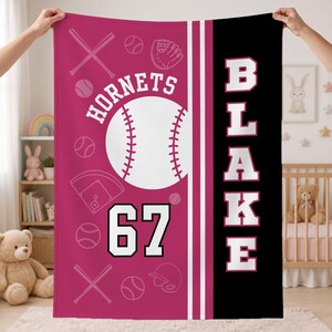 May include: A rectangular fleece blanket featuring a magenta left side with white baseball-themed graphics, including bats, balls, a glove, a helmet, and a home plate. The text reads HORNETS and 67. A black right side features the vertical text BLAKE.