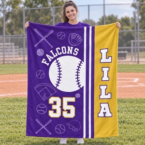 May include: A purple and gold fleece blanket featuring a large white baseball graphic, the text "FALCONS" in an arc, the number "35", and the name "LILA" vertically. The design includes white line art of baseball bats, gloves, and balls.