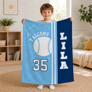 May include: A light blue and dark blue fleece blanket featuring a baseball theme with the text FALCONS, the number 35, and the name LILA. The design includes white baseballs, bats, and gloves on a light blue background with a dark blue side panel.