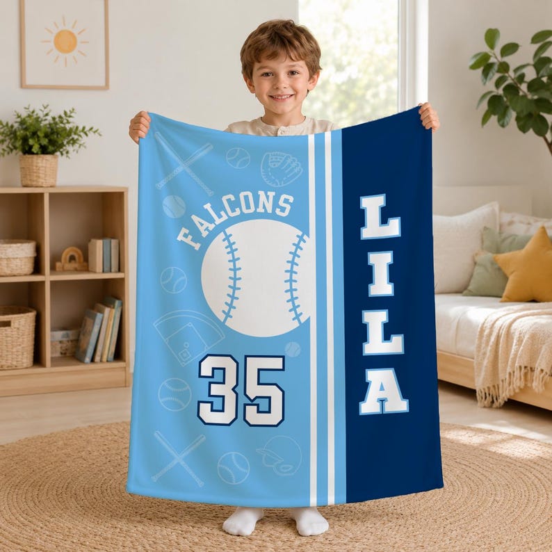 May include: A light blue and dark blue fleece blanket featuring a baseball theme with the text FALCONS, the number 35, and the name LILA. The design includes white baseballs, bats, and gloves on a light blue background with a dark blue side panel.