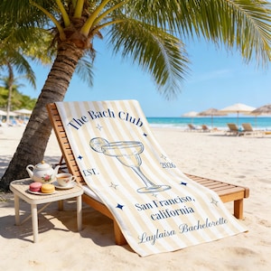 May include: A beach towel with vertical cream and yellow stripes, draped over a wooden beach chair. The towel features a margarita illustration and text that reads "The Bach Club", "San Francisco, California", and a name.