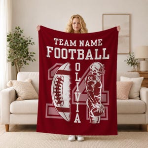 May include: A deep red fleece blanket featuring the white text TEAM NAME, FOOTBALL, and OLIVIA. The design includes a large white football graphic, a stylized football player in uniform, and a large number 14 in the background.