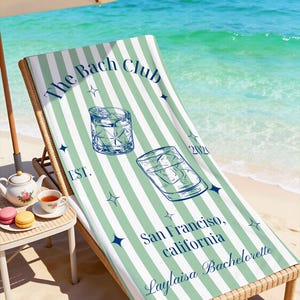 May include: A beach towel with a green and white striped pattern is draped over a wicker beach chair. The towel features the text "The Bach Club" and "San Francisco, California, Laylaisa Bachelorette". Two cocktail illustrations are also on the towel.