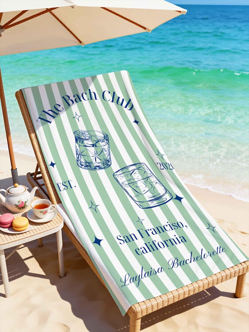 May include: A beach towel with a green and white striped pattern is draped over a wicker beach chair. The towel features the text "The Bach Club" and "San Francisco, California, Laylaisa Bachelorette". Two cocktail illustrations are also on the towel.