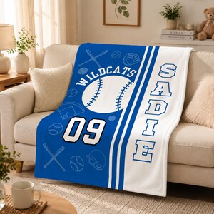 May include: A soft fleece blanket featuring a blue and white baseball theme. The design includes a large baseball, crossed bats, a glove, and the text Wildcats, 09, and the name Sadie in bold blue block lettering on a white vertical stripe.