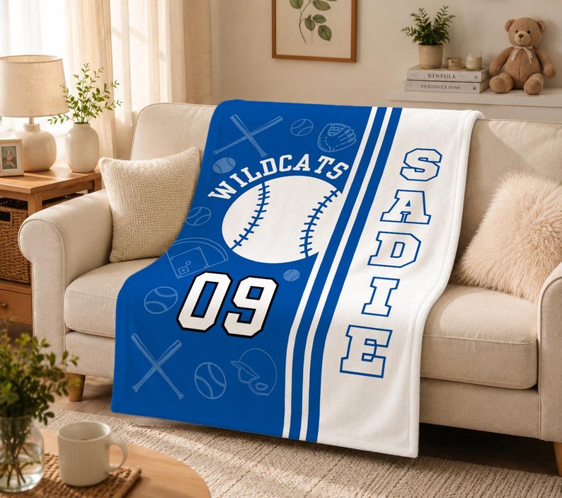 May include: A soft fleece blanket featuring a blue and white baseball theme. The design includes a large baseball, crossed bats, a glove, and the text Wildcats, 09, and the name Sadie in bold blue block lettering on a white vertical stripe.