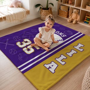 May include: A rectangular fleece blanket featuring a purple and gold color scheme with white baseball-themed graphics including bats, balls, and a glove. The blanket displays the number 35 and the name HILLA in large white block lettering.