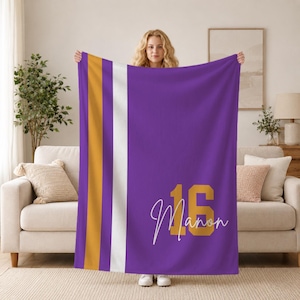 May include: A purple fleece blanket featuring a vertical gold and white stripe pattern on the left side. The blanket displays the large gold number 16 and the name Manon written in a white cursive script font on the lower right side.
