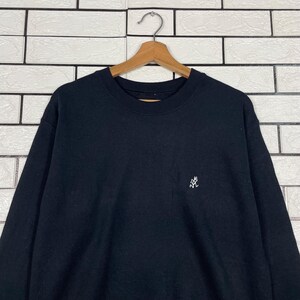 May include: Black crew neck sweatshirt with a small white embroidered logo on the chest. The sweatshirt is hanging on a wooden hanger against a white brick wall.
