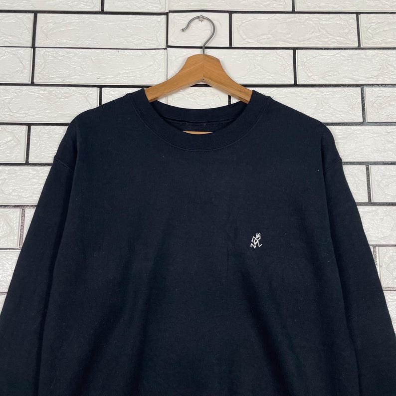 May include: Black crew neck sweatshirt with a small white embroidered logo on the chest. The sweatshirt is hanging on a wooden hanger against a white brick wall.
