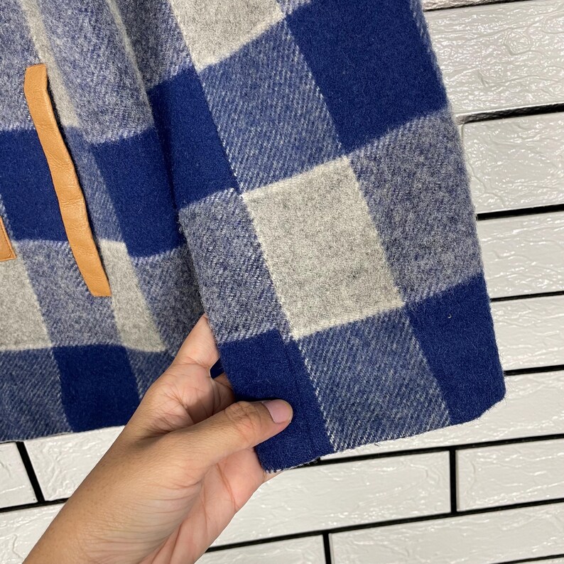 May include: Close-up of a blue, grey, and white plaid wool jacket. The jacket has a leather accent and a folded cuff. The fabric is thick and textured, with a classic check pattern. The jacket is a medium weight.