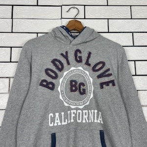 K&ouml;nnte beinhalten: Grauer Kapuzenpullover mit dem navyblauen Schriftzug "BODY GLOVE" in einem Bogen &uuml;ber der Brust. Darunter steht das Wort "CALIFORNIA" in Wei&szlig;. Der Pullover hat eine Kapuze mit Kordelzug und eine K&auml;ngurutasche.
