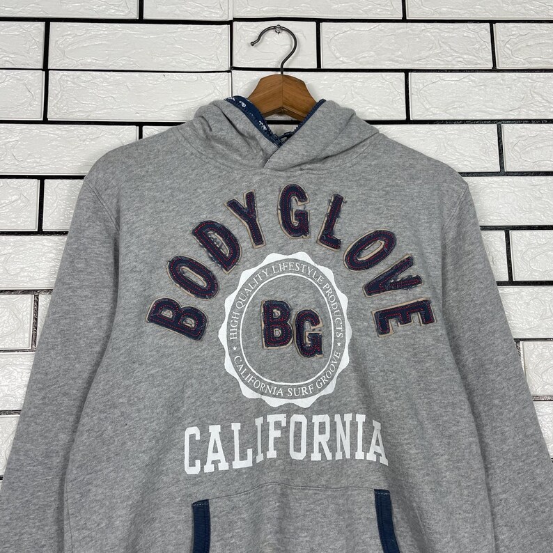 K&ouml;nnte beinhalten: Grauer Kapuzenpullover mit dem navyblauen Schriftzug "BODY GLOVE" in einem Bogen &uuml;ber der Brust. Darunter steht das Wort "CALIFORNIA" in Wei&szlig;. Der Pullover hat eine Kapuze mit Kordelzug und eine K&auml;ngurutasche.