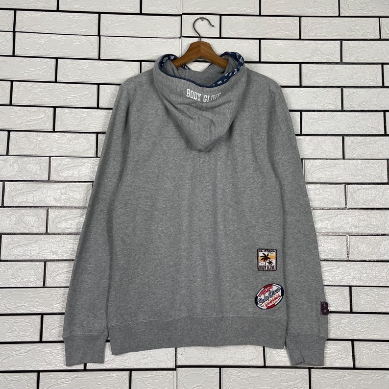 K&ouml;nnte beinhalten: Ein grauer Body Glove Hoodie mit Kapuze mit Kordelzug und blauem Musterfutter. Auf der R&uuml;ckseite befinden sich gestickte Aufn&auml;her mit Palmen und Text. Der Hoodie h&auml;ngt an einem Holzb&uuml;gel.