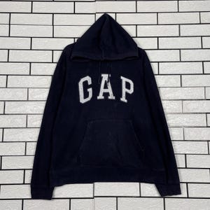 Könnte beinhalten: Ein marineblauer Kapuzenpullover mit dem weißen Text "GAP" auf der Brust. Der Pullover hat eine Vordertasche und eine Kapuze mit Kordelzug. Das Kleidungsstück ist aus einem weichen, bequemen Material gefertigt.