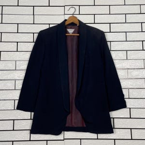 May include: A dark navy blazer with a shawl collar, hanging on a wooden hanger. The interior lining is a deep burgundy color. The blazer is displayed against a white brick wall.