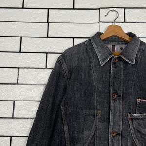 May include: A dark grey denim jacket with a button-up front, collar, and pockets. The jacket is hanging on a wooden hanger against a white brick wall. The denim has a worn appearance.