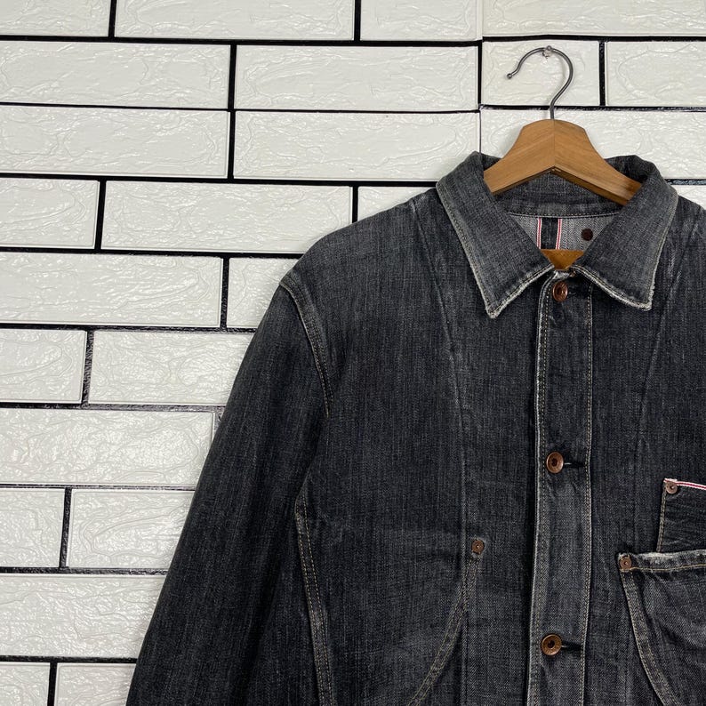 May include: A dark grey denim jacket with a button-up front, collar, and pockets. The jacket is hanging on a wooden hanger against a white brick wall. The denim has a worn appearance.