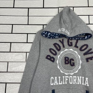 K&ouml;nnte beinhalten: Grauer Kapuzenpullover mit dem Text "BODY GLOVE" in einem kreisf&ouml;rmigen Design. Die Kapuze hat eine Bord&uuml;re mit Paisley-Muster und den Text "BODY GLOVE" aufgedruckt. Der Pullover tr&auml;gt auch den Text "CALIFORNIA".