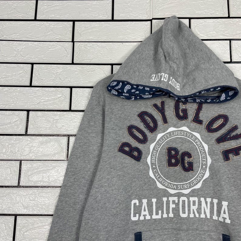K&ouml;nnte beinhalten: Grauer Kapuzenpullover mit dem Text "BODY GLOVE" in einem kreisf&ouml;rmigen Design. Die Kapuze hat eine Bord&uuml;re mit Paisley-Muster und den Text "BODY GLOVE" aufgedruckt. Der Pullover tr&auml;gt auch den Text "CALIFORNIA".