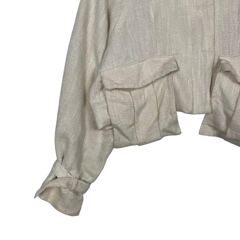 May include: Cream-colored cropped jacket with two front pockets. The jacket has long sleeves with a knotted detail at the wrist. The fabric has a subtle, textured appearance.