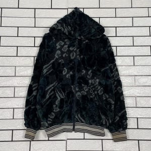 May include: A black and dark gray zip-up hoodie featuring a textured, plush faux fur finish with an abstract animal print pattern. The garment includes a hood, long sleeves, and ribbed cuffs and hem with horizontal stripes in a neutral tone.