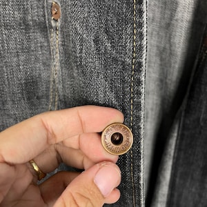 May include: Close-up of a dark grey denim jacket with a bronze button. The button is engraved with the word "EDWIN". The jacket's stitching is visible.