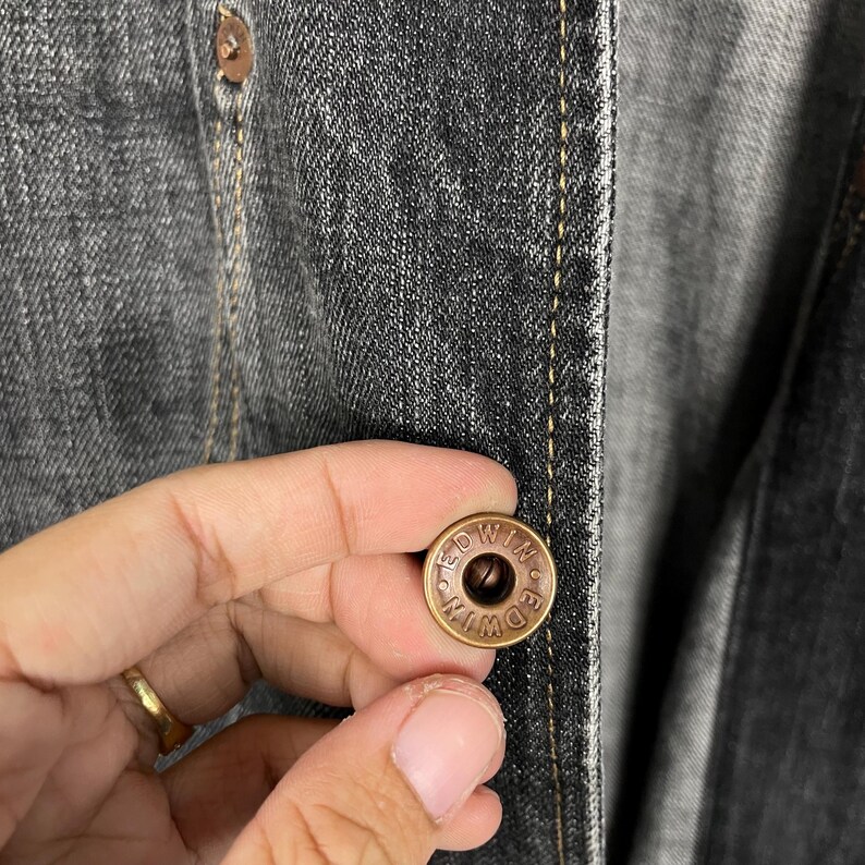 May include: Close-up of a dark grey denim jacket with a bronze button. The button is engraved with the word "EDWIN". The jacket's stitching is visible.