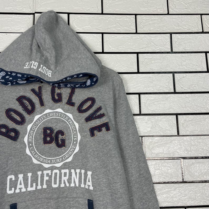 K&ouml;nnte beinhalten: Ein grauer Kapuzenpullover mit dem Text "BODY GLOVE" in dunkelvioletten Buchstaben. Der Pullover hat ein wei&szlig;es kreisf&ouml;rmiges Logo mit "BG" innen und "CALIFORNIA SURF GROOVE" darum. Die Kapuze hat eine marineblaue gemusterte Bord&uuml;re.