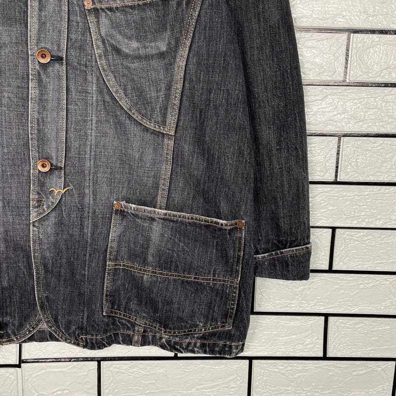 May include: A dark grey denim jacket with a button-up front, two front pockets, and a unique seam design. The jacket has a classic collar and long sleeves. The buttons are a light brown colour.