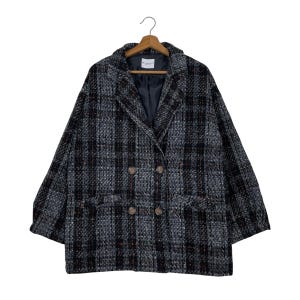 Pode incluir: Um blazer oversized de abotoamento duplo com um padrão xadrez cinza, preto e marrom. O casaco apresenta uma gola entalhada, dois bolsos frontais e quatro botões marrons. Pendurado em um cabide de madeira.