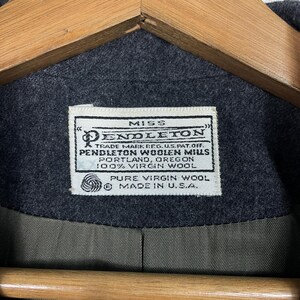May include: A close-up of a dark grey wool coat featuring a label that reads "MISS PENDLETON". The label also states "PENDLETON WOOLEN MILLS, PORTLAND, OREGON, 100% VIRGIN WOOL". The coat is made in the U.S.A.