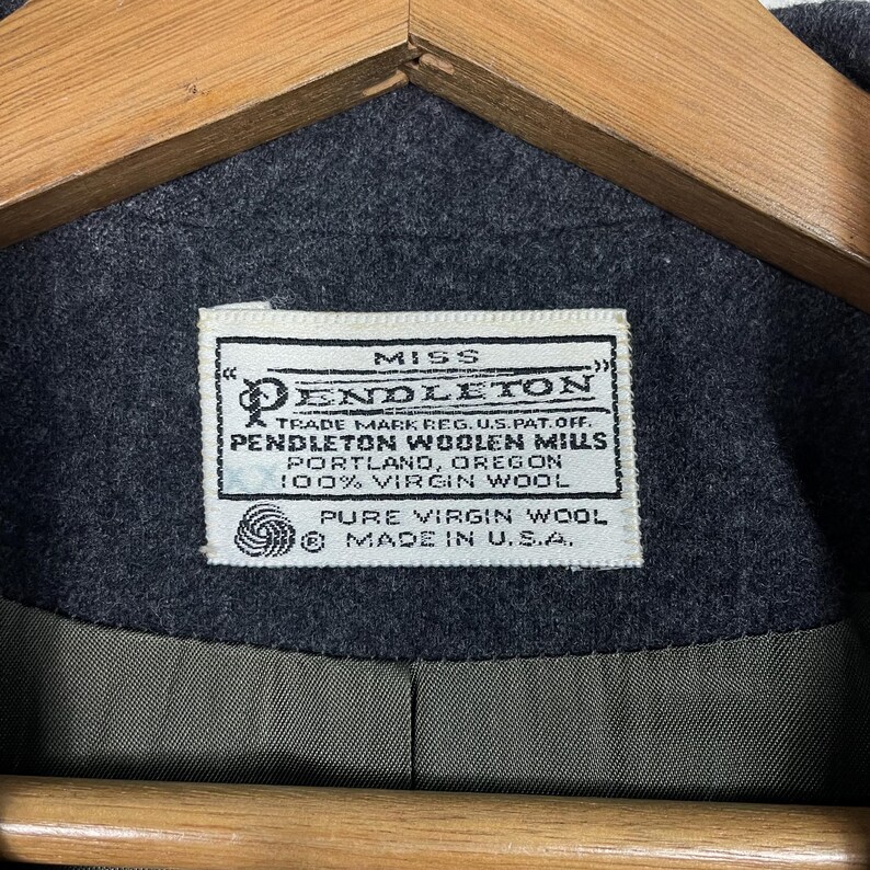 May include: A close-up of a dark grey wool coat featuring a label that reads "MISS PENDLETON". The label also states "PENDLETON WOOLEN MILLS, PORTLAND, OREGON, 100% VIRGIN WOOL". The coat is made in the U.S.A.