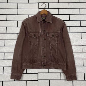 Op de afbeelding: Een bruine spijkerjas met een knoopsluiting, twee borstzakken en twee zijzakken. De jas hangt aan een houten hanger tegen een witte bakstenen muur.